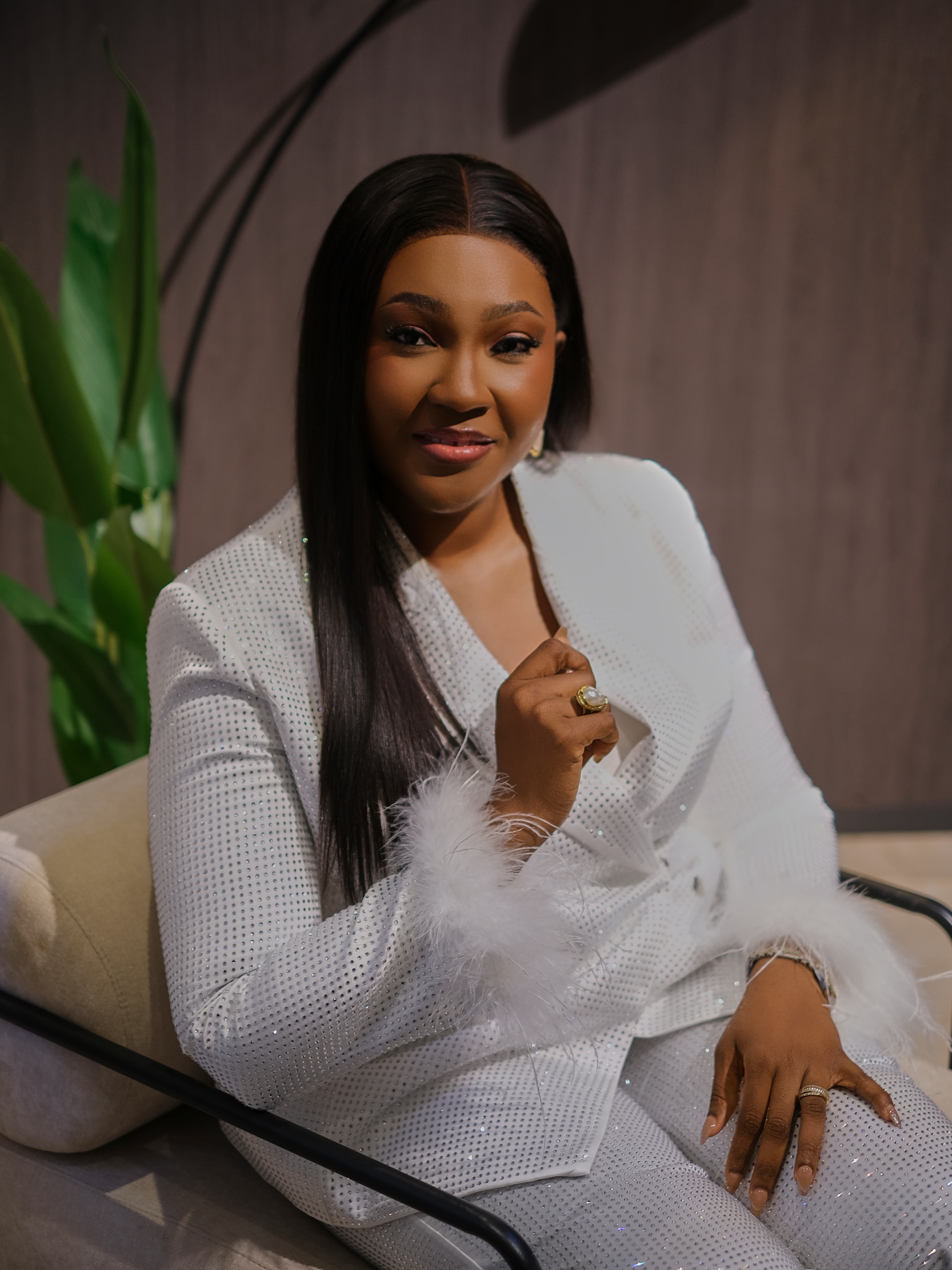Pastor Adeola Ajani, seated, warmly smiling, in a white sequined suit.